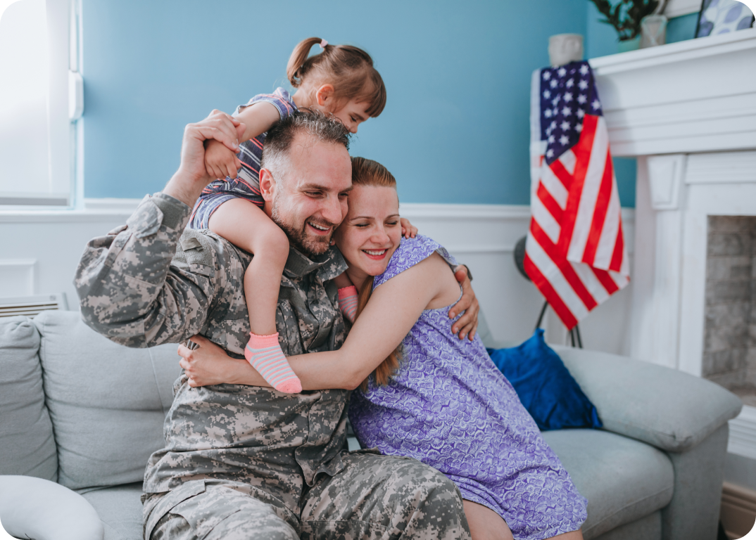 A man in military fatigues holding his wife and daughter while sitting on a couch in a living room.