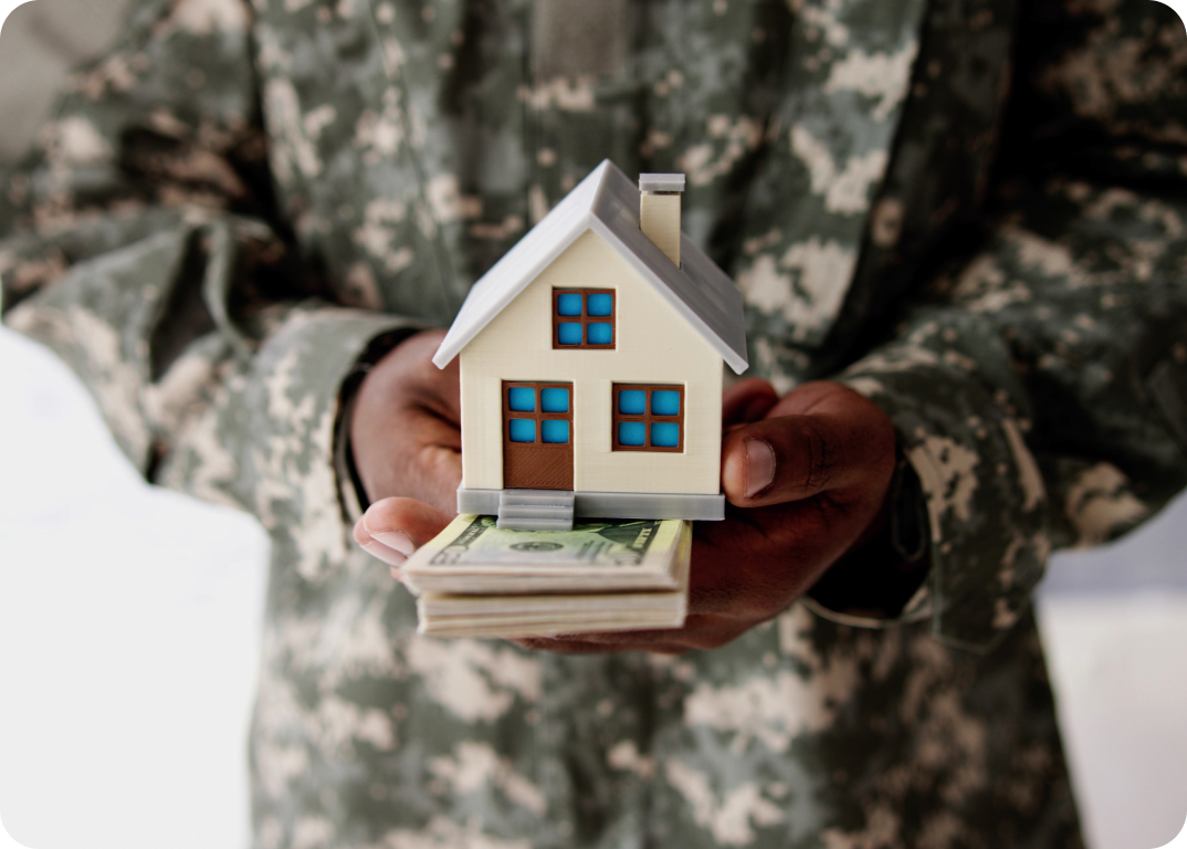 A person holds a miniature model of a house with a stack of bills below it.